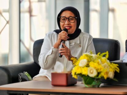 Woman wearing a black hijab and glasses speaks into a microphone during a panel with yellow flowers on the table nearby